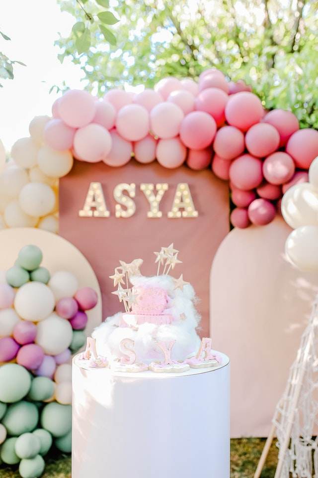 Decorative birthday party display for "Asya" featuring pink and purple balloons and a themed cake under a balloon arch at an outdoor event.