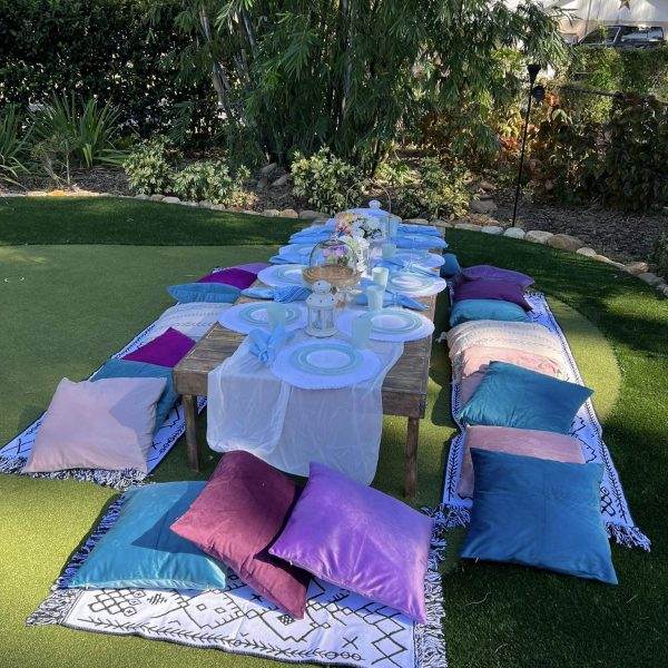 An outdoor dining setup on a grassy area with a long table covered with a white cloth, surrounded by colorful pillows for seating, and elegant table settings under palm trees.