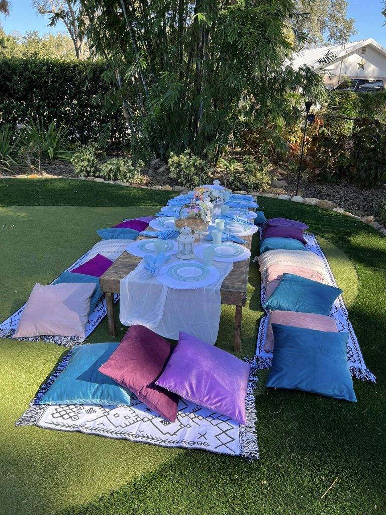Outdoor dining setup with a long table, blue and white tableware, surrounded by colorful cushions on carpets, set on artificial grass, with palm trees in the background, offering an upscale outdoor dining experience.