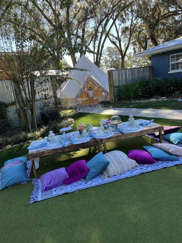 Outdoor children's party setup with a luxury picnic, including a teepee, a low table set with tea party items, and colorful cushions on artificial grass.