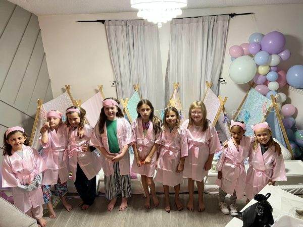 A group of young girls, all wearing pink robes and matching headbands, are smiling and posing together at a slumber party, surrounded by 314436325_1519558038468382_8873227985688846232_n and decorations.