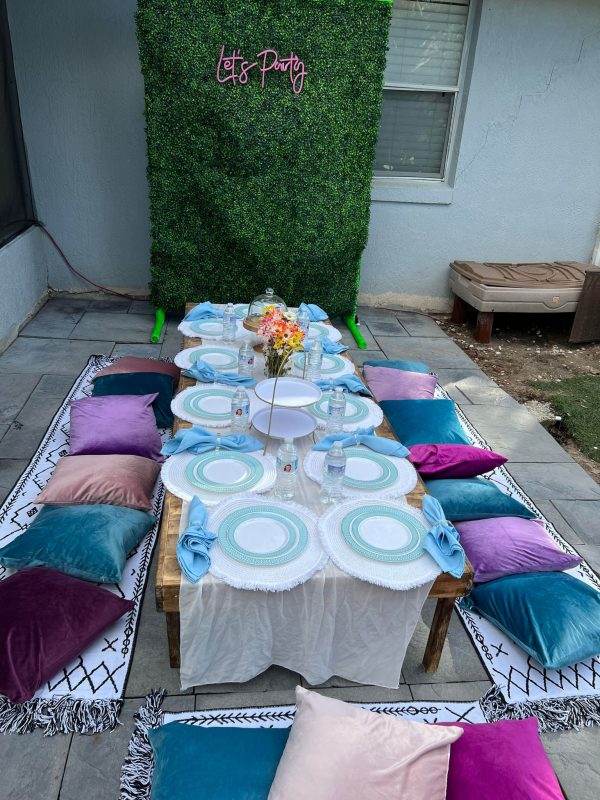 An outdoor party setting with a 315520845_1469721590105982_9118067481530006582_n circular table, surrounded by cushions in pink, purple, and teal, under a "let's party" sign on a green hedge wall.