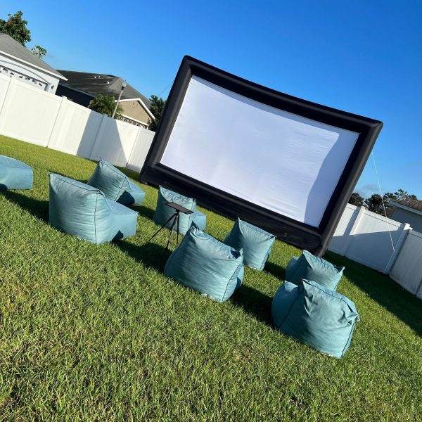An outdoor movie setup with a large inflatable screen and several 315530724_8394359350635245_1804058737834671934_n bean bag chairs on a green lawn, under clear blue skies.