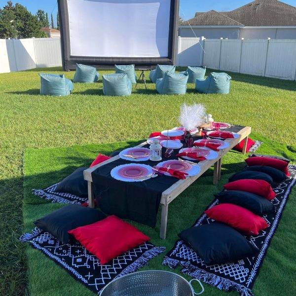 Outdoor glamping and movie rental night setting with a large inflatable screen, a low wooden table set for a luxury picnic, red and black cushions on the grass, and a metal bucket in the foreground.