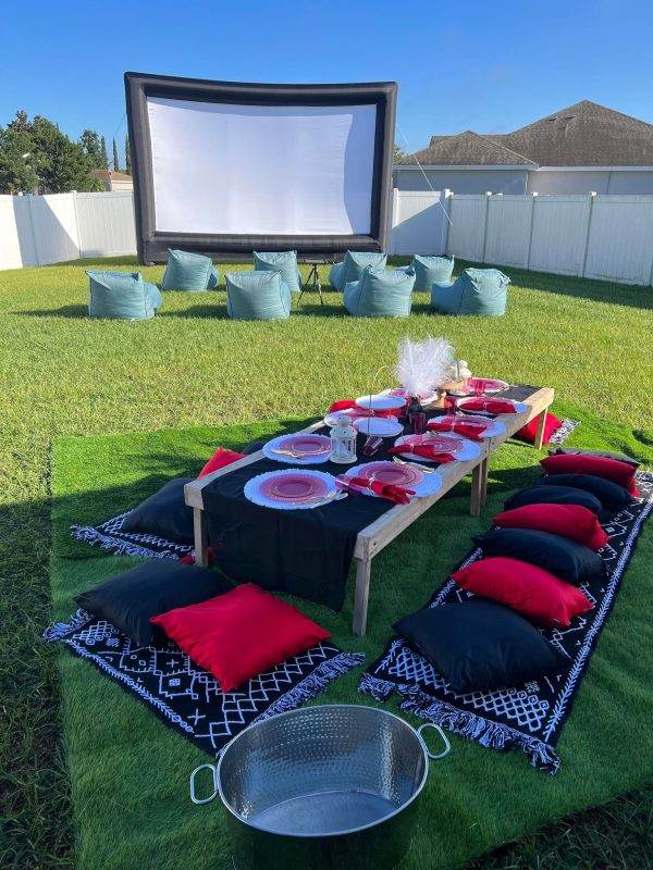 Outdoor glamping and movie rental night setting with a large inflatable screen, a low wooden table set for a luxury picnic, red and black cushions on the grass, and a metal bucket in the foreground.