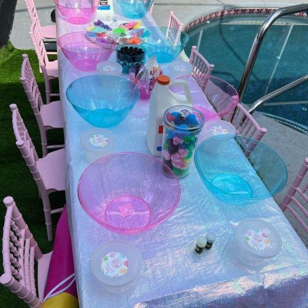 A colorful outdoor slime party setup with a table set up with blue and pink bowls and plates perfect for a glamping experience, adorned with pink chairs, large bowls, cups, and craft supplies, next to a swimming pool.