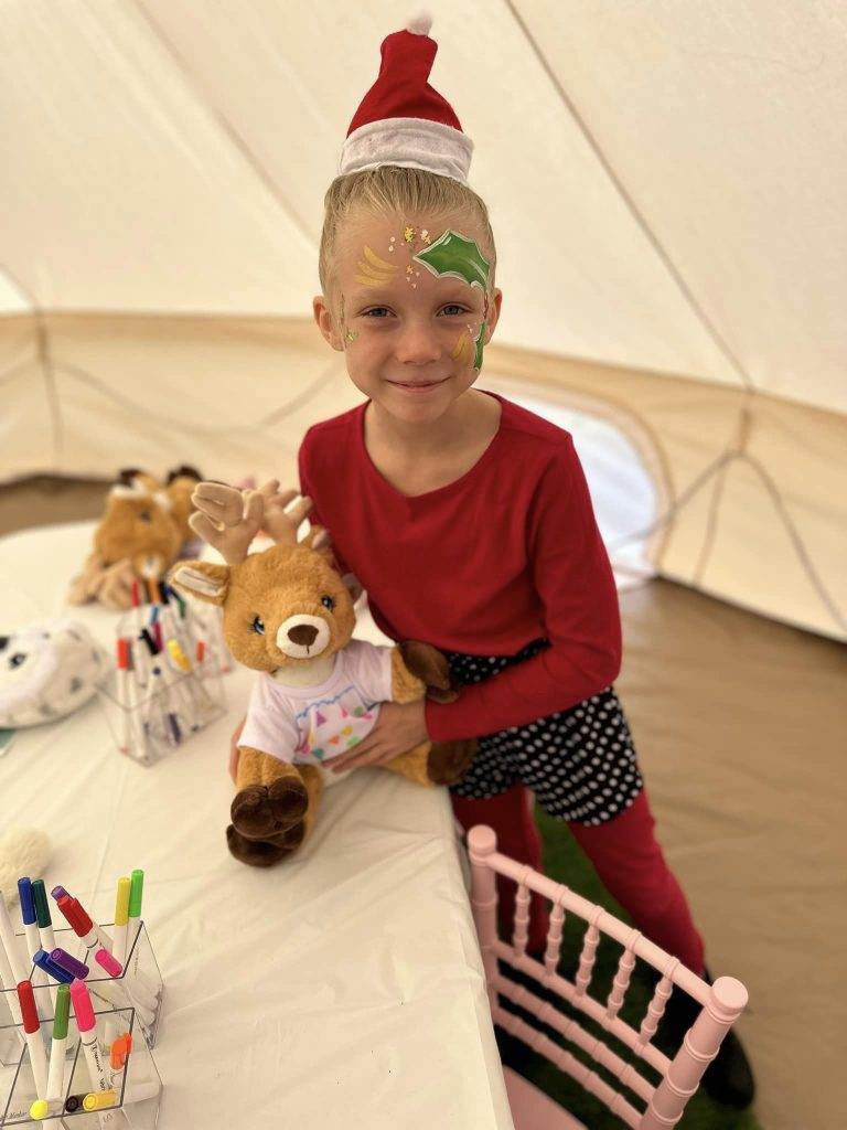 A young child with face paint and a festive hat smiles while holding a Bell Tent, sitting at a table with various toys inside a light-colored tent.