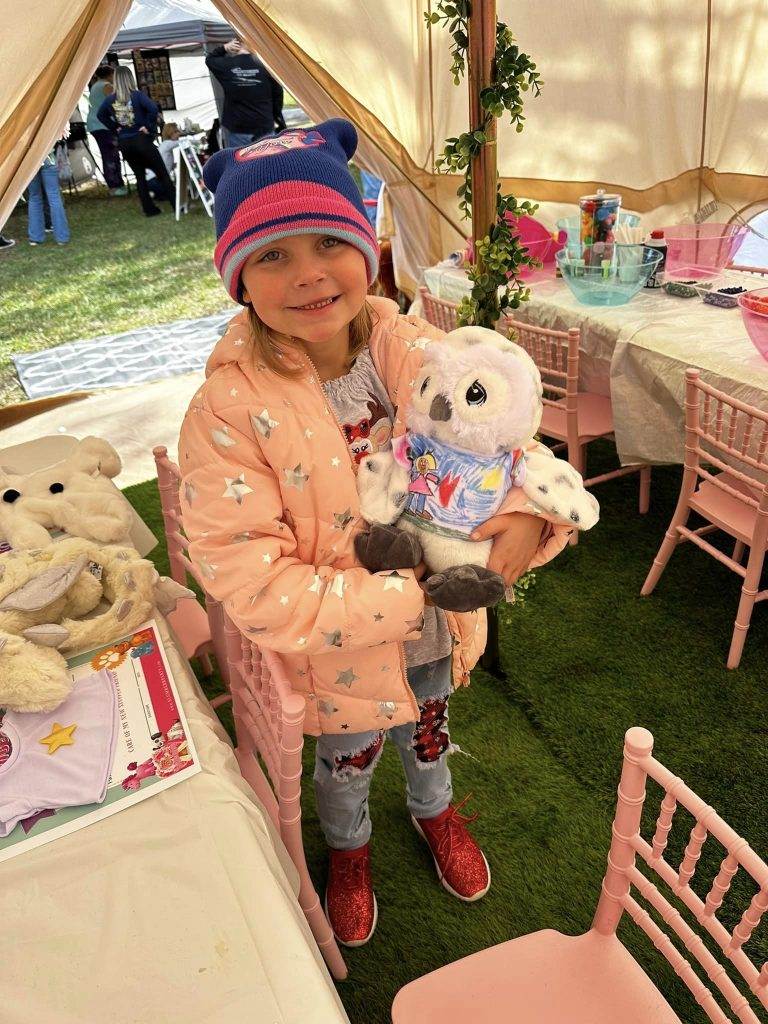 A young girl in a pink starry jacket and blue hat, smiling and holding a plush Teddy bear, stands in a teepee tent decorated with pink chairs and tables.
