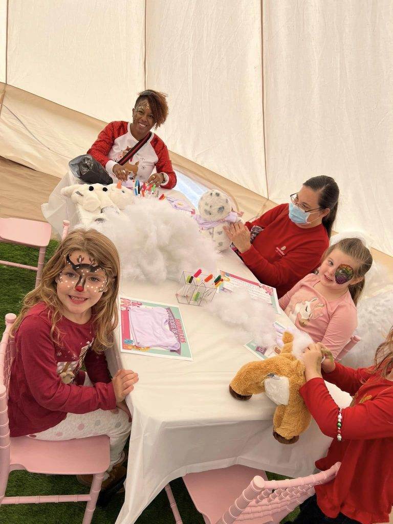 A lively craft session inside a tent with a group of children sitting at a Lakeland table in a Bell Tent and making stuffed animals. Everyone smiles, surrounded by fluffy white stuffing and colorful markers.