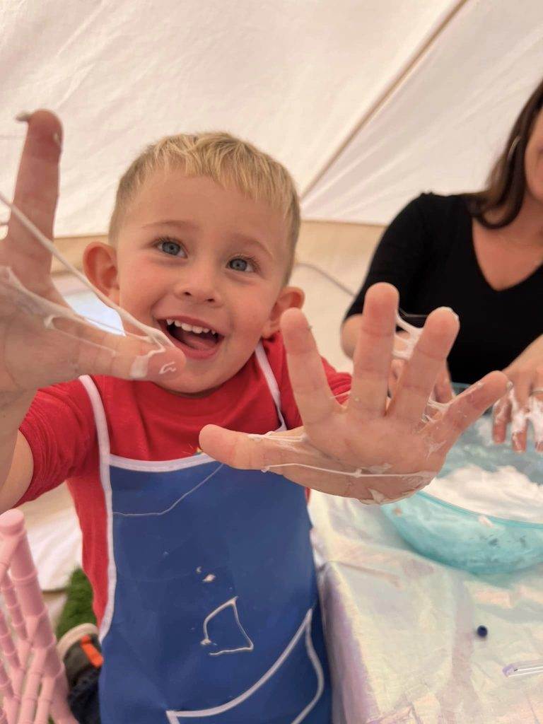 A joyful young boy with blue eyes and short blond hair, wearing a blue apron, shows his hands covered in white paint under a Lakeland party. he is smiling broadly, indicating he is having fun.