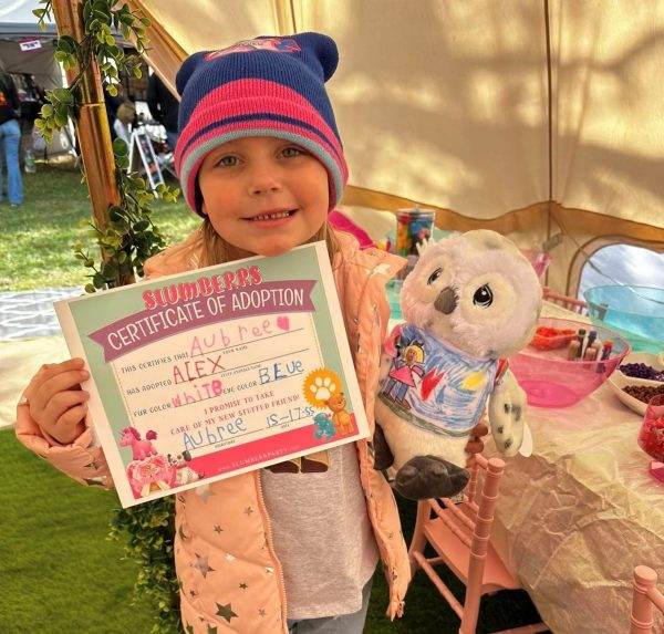 Young girl holding a certificate of adoption and a stuffed toy from a Build A Bear Party at an outdoor event. She wears a pink hat and a smile.