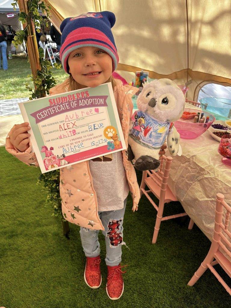 A little girl holding a "certificate of adoption" for a stuffed owl in front of a teepee tent. The child wears a winter hat, jacket, and red shoes, standing in a tent with a table and chairs nearby.