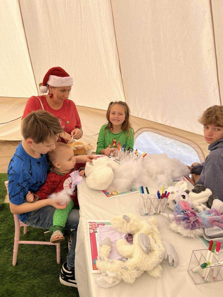 A group of people sitting at a Bell Tent in Lakeland, crafting stuffed toys. a woman in a santa hat assists while two boys and a girl engage in activities, surrounded by fluffy white materials and colorful tools.