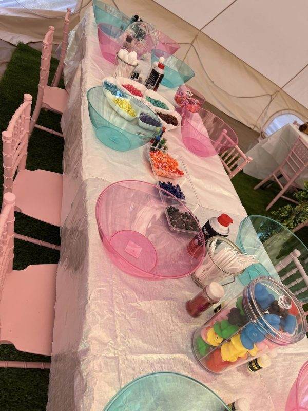A party tent set up with pink and blue bowls filled with various colorful candies and toppings, and white chairs aligned along its side for a glamping themed event.