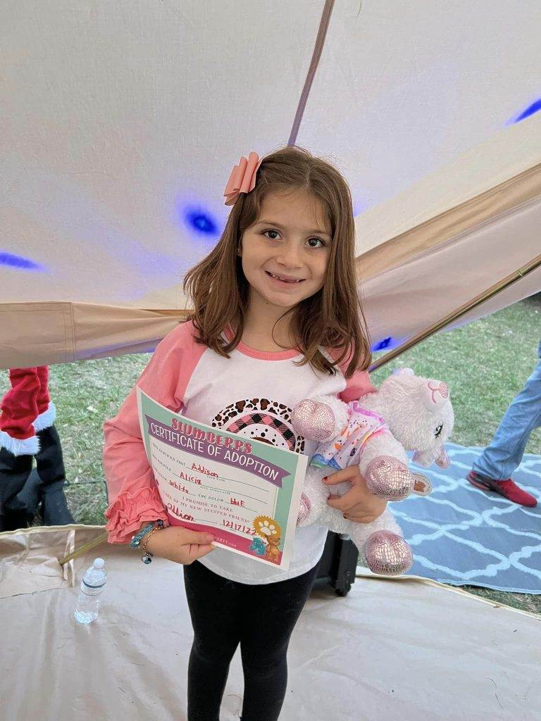 A young girl holding a My Little Pony plush toy and a "certificate of adoption." She wears a pink and white shirt with a ribbon in her hair, standing under a white tent.