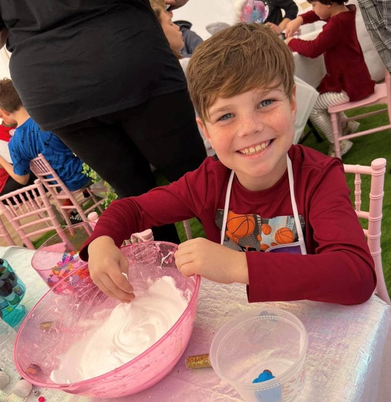 A young boy with a joyful expression mixes a large bowl of pink slime at a slime party, wearing a basketball-themed apron.