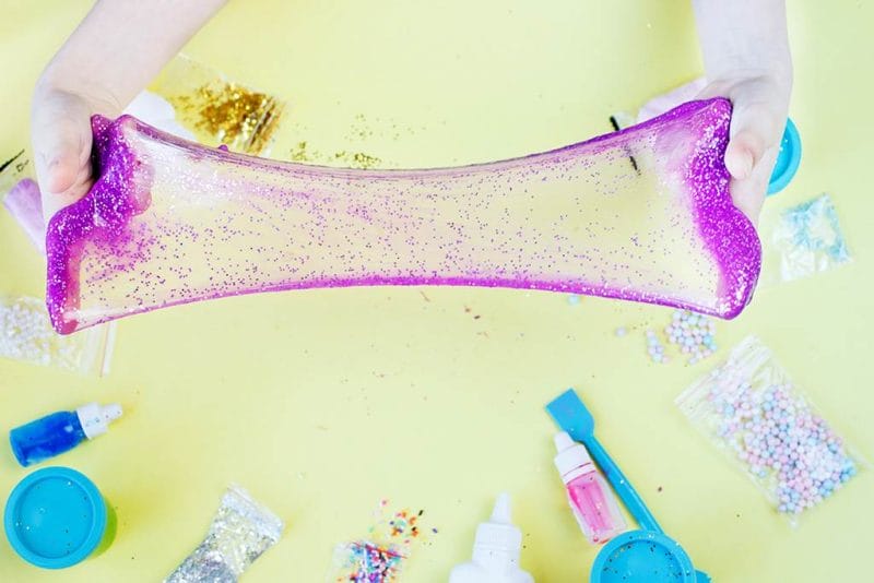 Hands holding a stretched piece of pink glittery slime over a yellow table scattered with craft supplies, ready for the perfect slime party.
