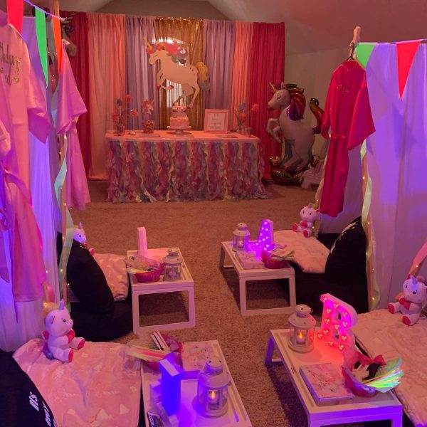 Indoor unicorn-themed birthday party setup with pink and rainbow decorations, small tables with cushions from a teepee company, and unicorn figures.