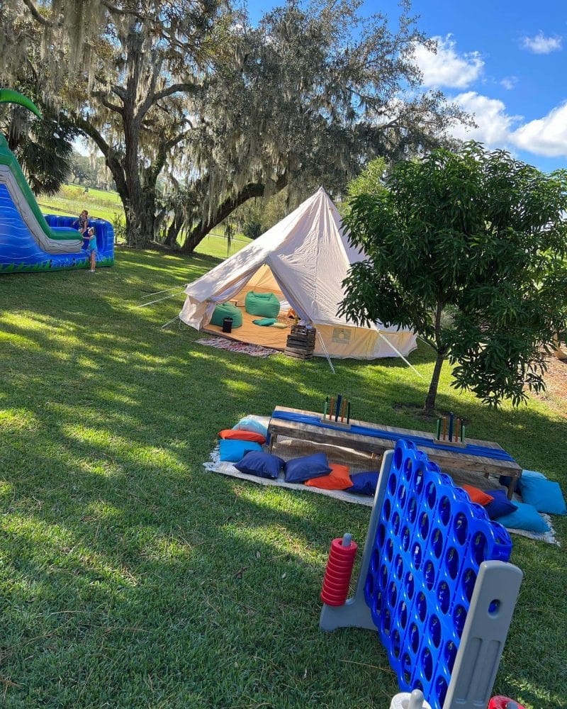 A spacious outdoor area featuring a large tent, inflatable slides, and a giant connect four game on a sunny day with scattered trees, perfect for kids entertainment at weddings.