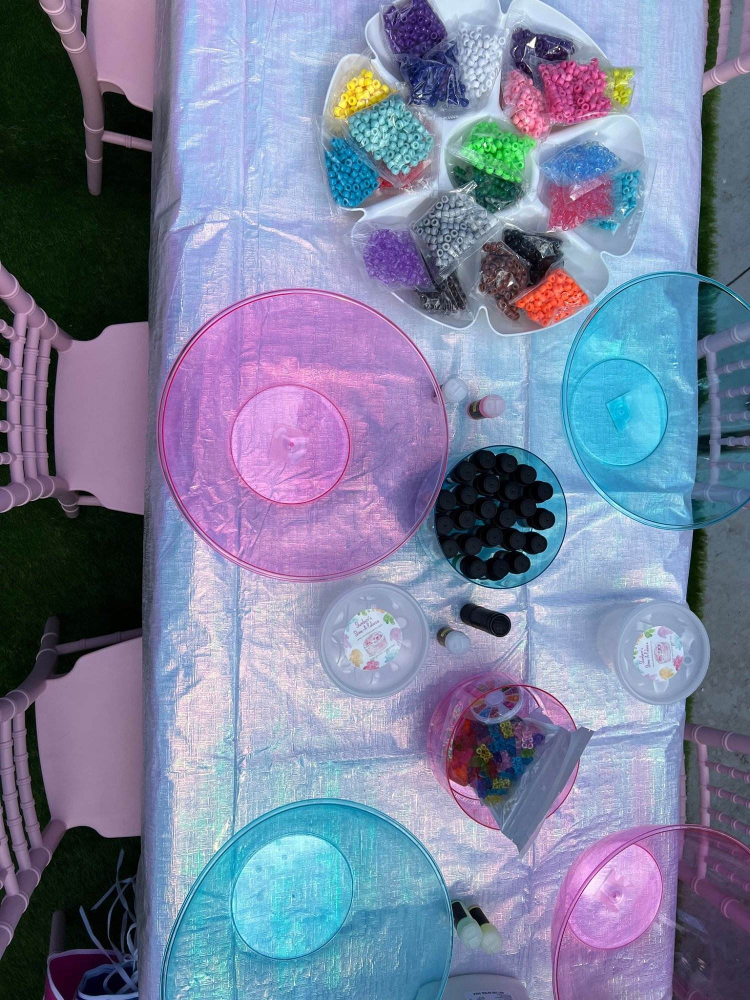 Overhead view of a crafting table designed to entertain kids during a wedding, with colorful beads in assorted containers and empty plastic bowls on a pink tablecloth.