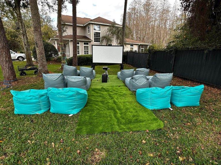 Outdoor home cinema setup with blue bean bags on green artificial turf, perfect for event planning, features a large screen, and is surrounded by trees and a wooden fence.