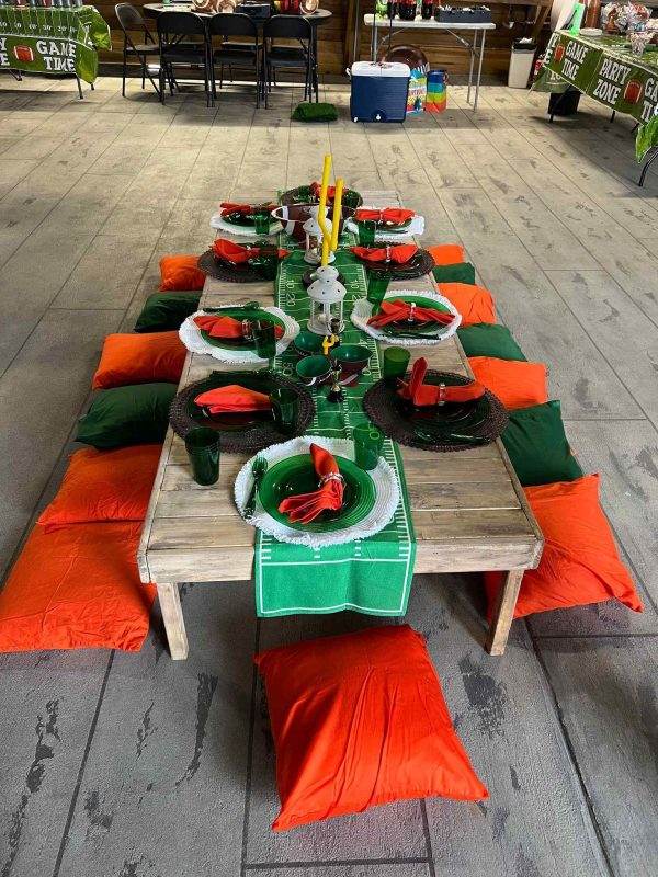 A festive outdoor dining setup with a Teepee set up for a Lakeland football game, offering a unique Glamping experience flanked by orange and green pillows on the floor, set with black plates, red napkins, and a green runner under game time banners.