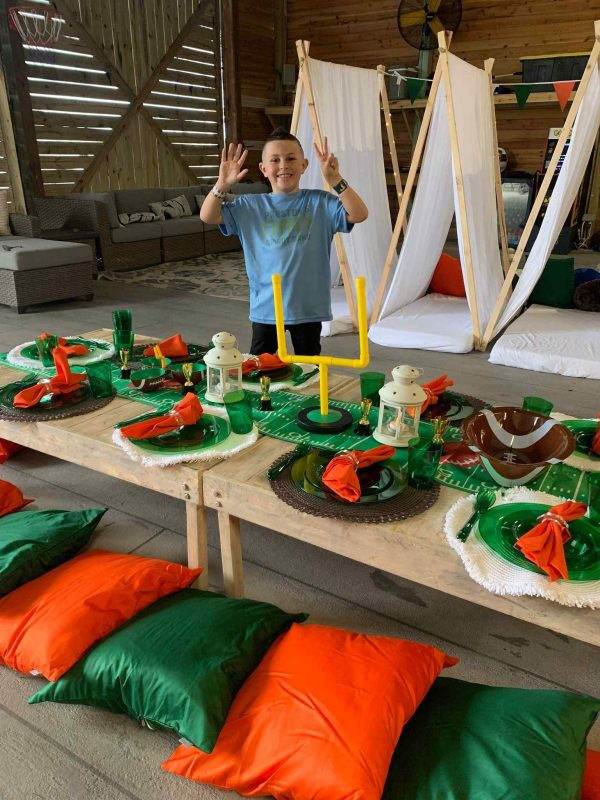 A boy is posing for a picture in front of a Teepee at a festive party table decorated in orange and green, with football-themed plates and centerpieces, surrounded by hanging white tents and floor cushions.