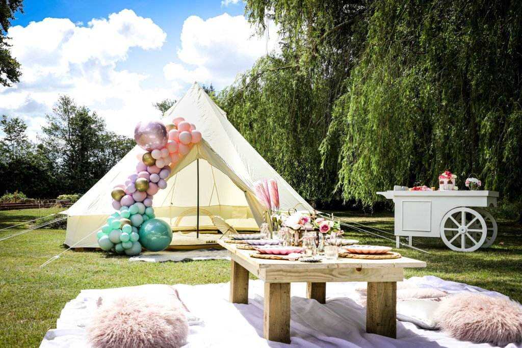 Outdoor wedding entertainment setup with a canvas tent adorned with colorful balloons, a wooden table with a feast, and a white cart under a clear sky designed to entertain kids.