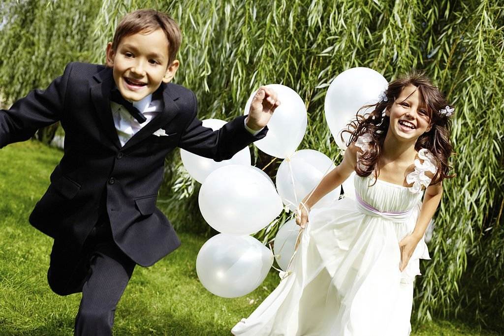 A boy and a girl, dressed in formal attire, joyfully run through a grassy area holding white balloons as part of the wedding entertainment.
