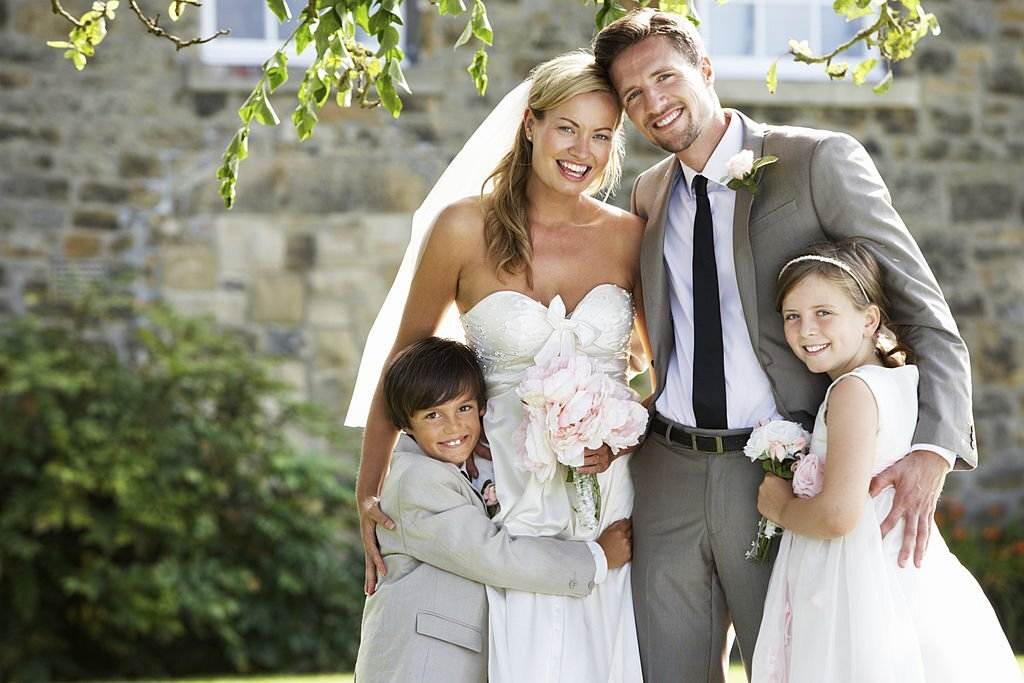A bride and groom smiling outdoors with two young kids, a boy and a girl, dressed in formal attire, standing in front of a stone building.