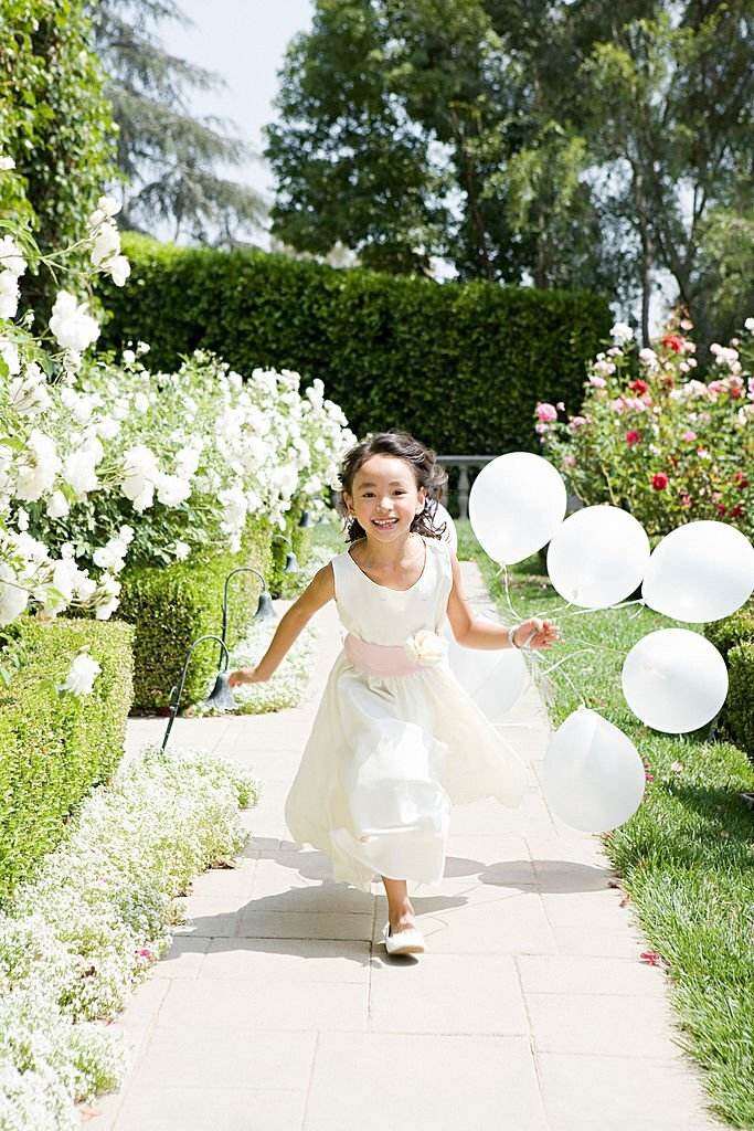 Young girl in a white dress running along a garden path, holding white balloons, smiling joyfully in a sunny, flower-lined setting during wedding activities.
