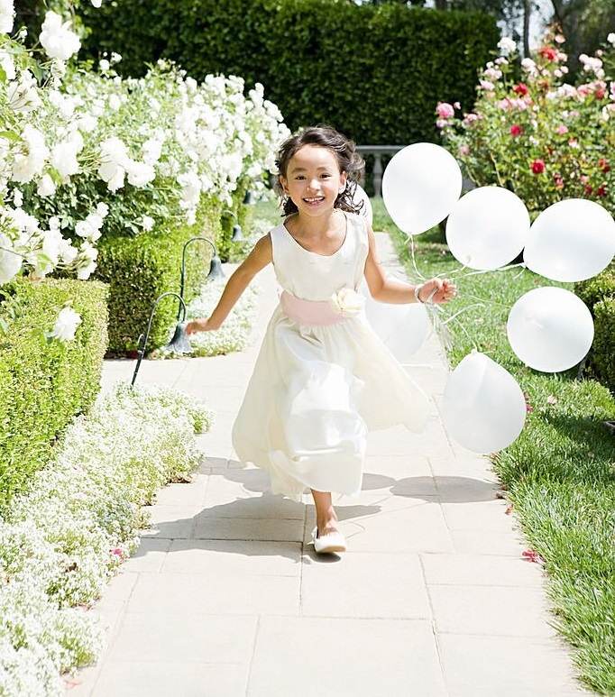 Young girl in a white dress joyfully running along a garden path holding white balloons and a lantern under a sunny sky during wedding activities.