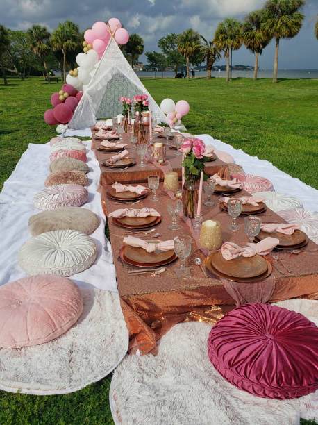 Outdoor dining setup with a long table covered in a copper cloth, surrounded by pink and white pillows under a white canopy, facing a lakeside view with grass and trees for a luxury picnic celebration.