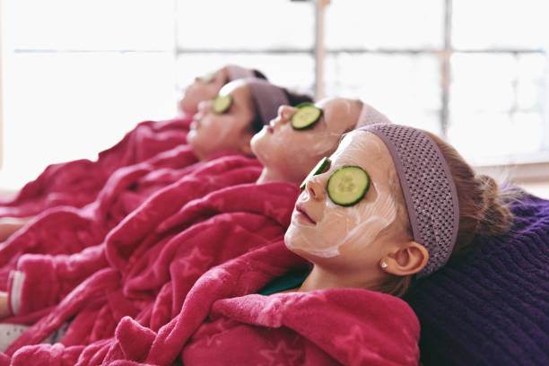 Three people lying down with facial masks and cucumber slices on their eyes, covered with pink blankets, relaxing in a home spa setting.