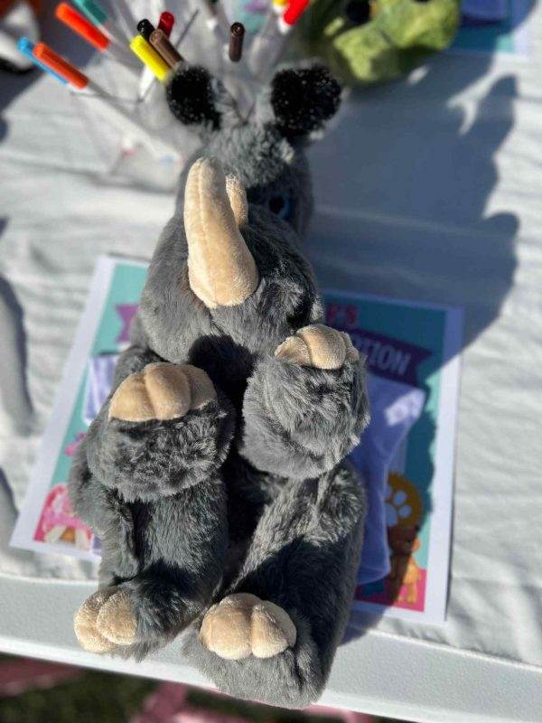 A stuffed rhino with large ears and eyes, sitting on a Lakeland table surrounded by crayons and coloring books, outdoors under bright sunlight at a Build a Bear party.