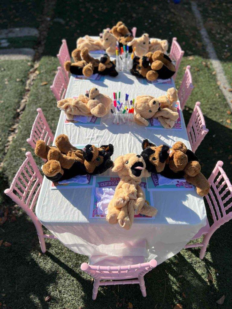 Teddy bears seated around two small pink tables set for a Build A Bear party outdoors, with coloring books and crayons.