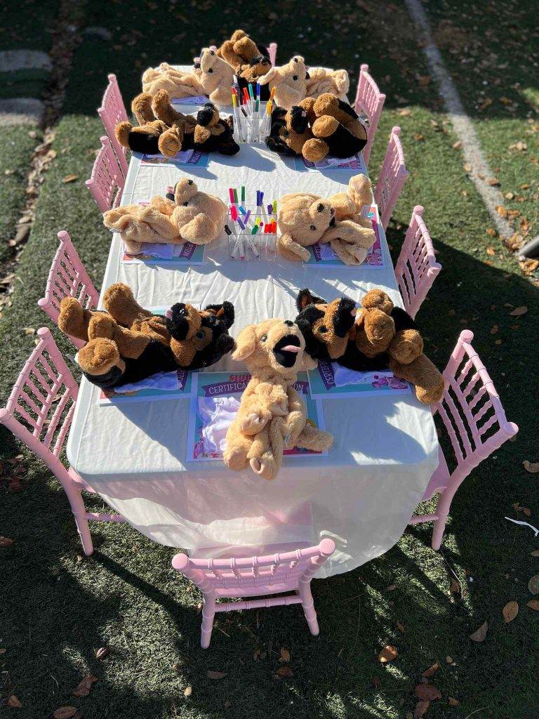 Outdoor children's Build A Bear party table set up with stuffed teddy bears seated around, under trees casting shadows, with straws and napkins ready.