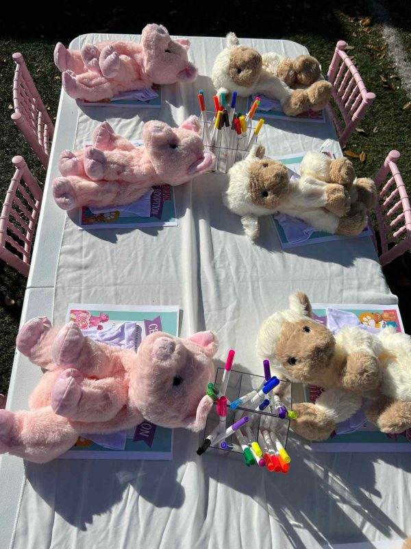 Stuffed animals, including custom bears, sitting at tables with coloring books and markers, set up outdoors for a children's activity.