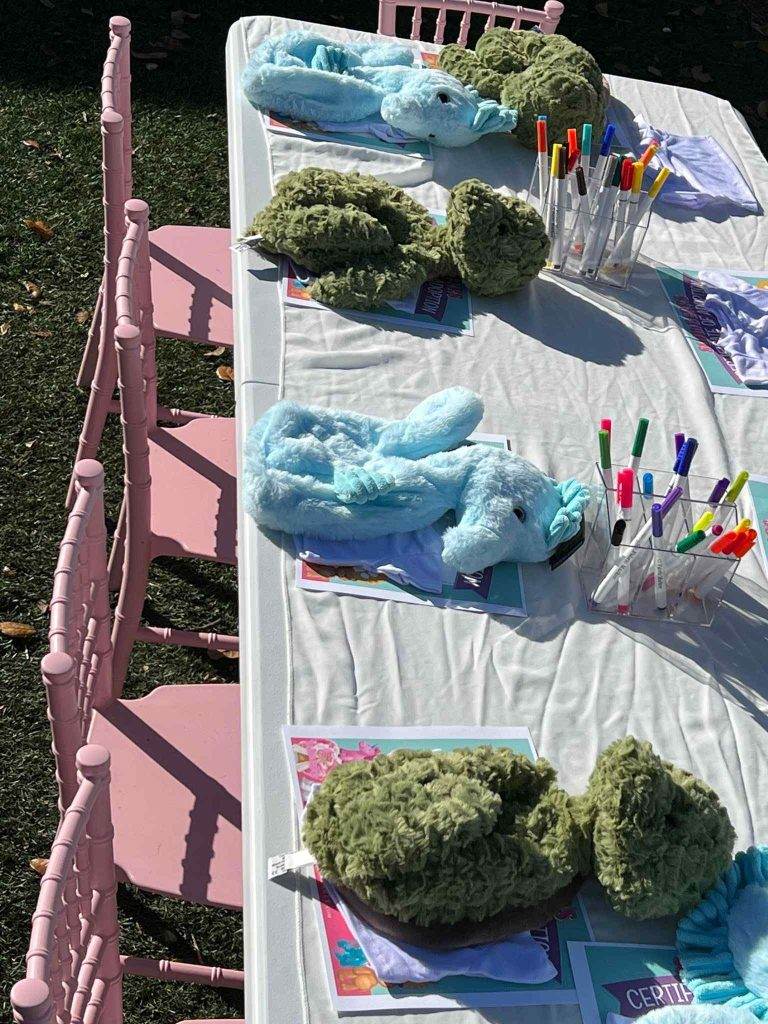 Table with green and blue plush crocodile toys from Build a Bear and coloring materials, set up outdoors with certificates on the table edge.