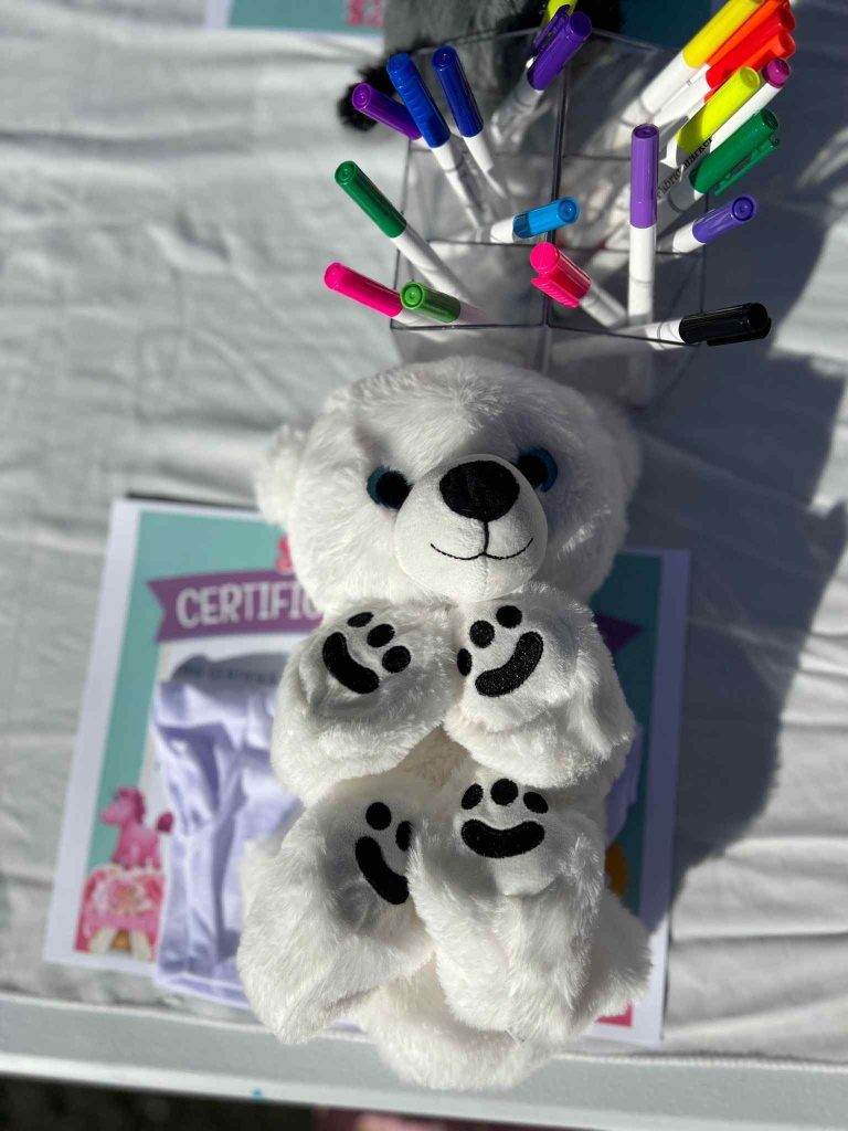 White teddy bear surrounded by colorful markers and a certificate on a table, set up for Build A Bear Party activities, in bright outdoor lighting.