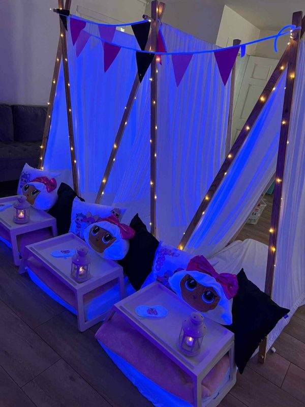Indoor sleepover setup with three cushioned floor beds under a canopy of blue lights and white drapes, adorned with purple and pink bunting. each bed has a plush unicorn pillow and a small wooden tray with a lantern.