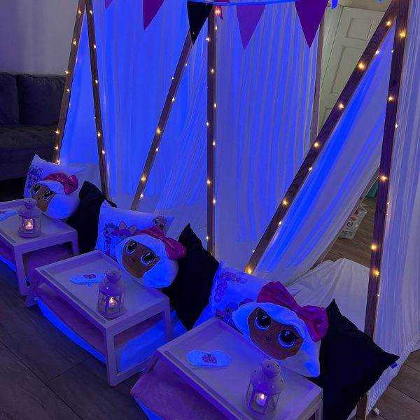 Indoor camping setup with party themes, including three beds under a white canopy, decorated with purple string lights and flags, each bed containing a plush toy and a serving tray.