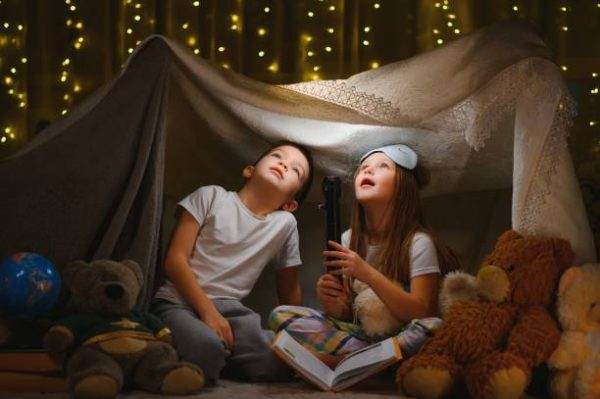 Two children, a boy and a girl, sitting in a blanket fort lit by fairy lights, looking up with wonder as the girl holds a flashlight during their slumber party.