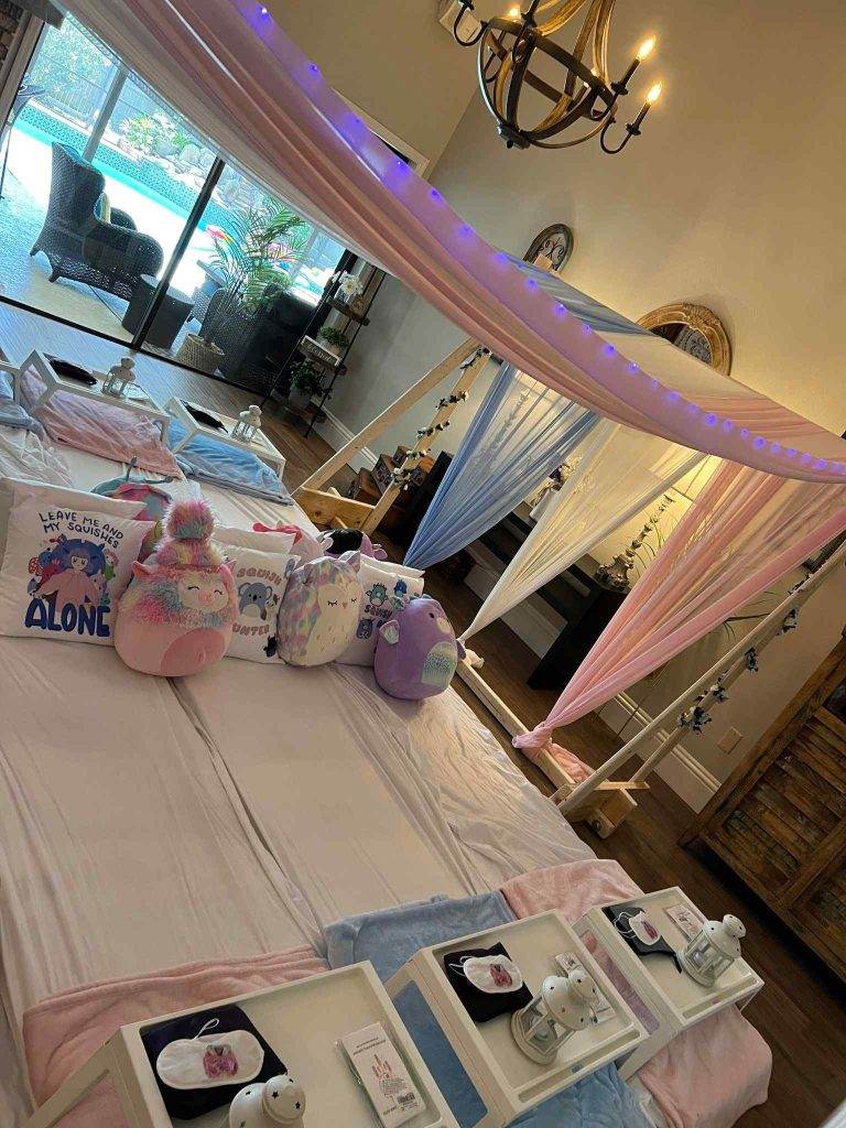 Interior room setup for a canopy slumber party with massage tables draped in pink and blue, plush slippers and robes placed on each, and string lights on the ceiling.