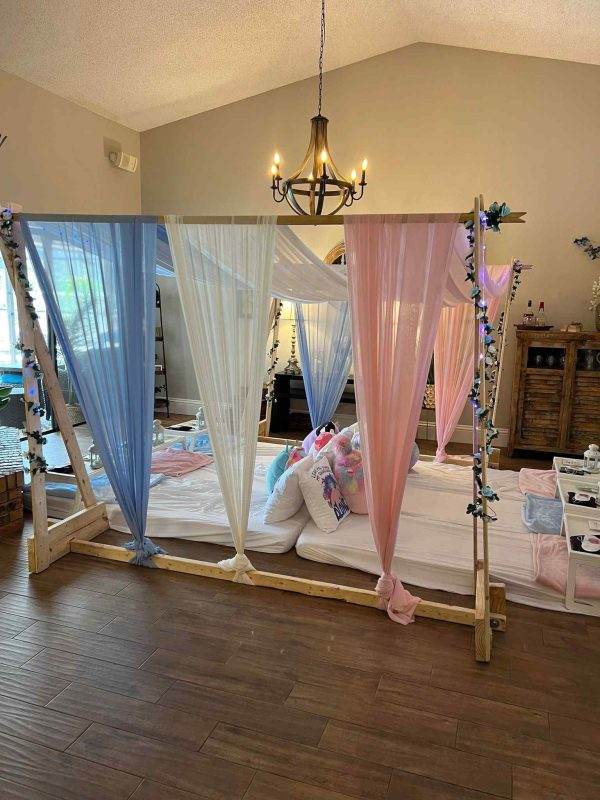 A wooden canopy bed draped with blue and pink sheer curtains, prepared for a kids sleepover party, in a room with a chandelier and decorative items.