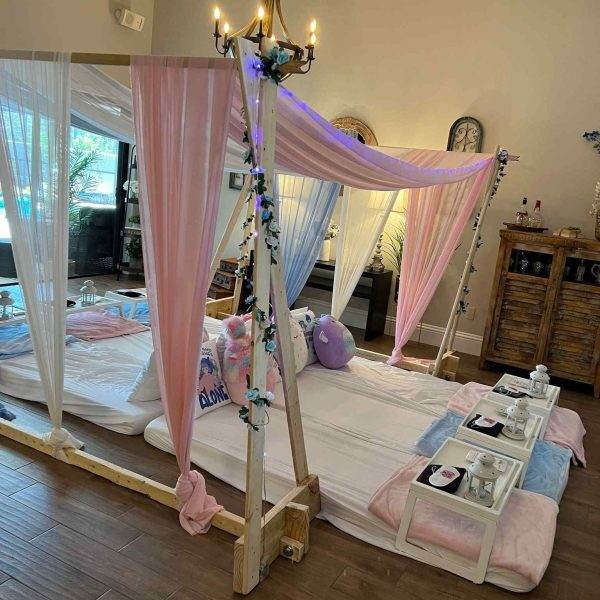 A cozy indoor bedroom setup featuring a pink and blue Bell Tent set up in a living room, adorned with light pink sheer drapes, fairy lights, and plush toys, creating a whimsical ambiance perfect for a slumber.
