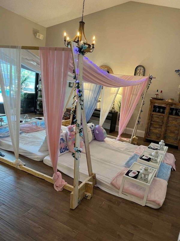 A cozy indoor bedroom setting with a canopied bed draped in pink curtains, adorned with fairy lights and ivy, next to a tray with spa essentials, perfect for a Canopy Slumber Party