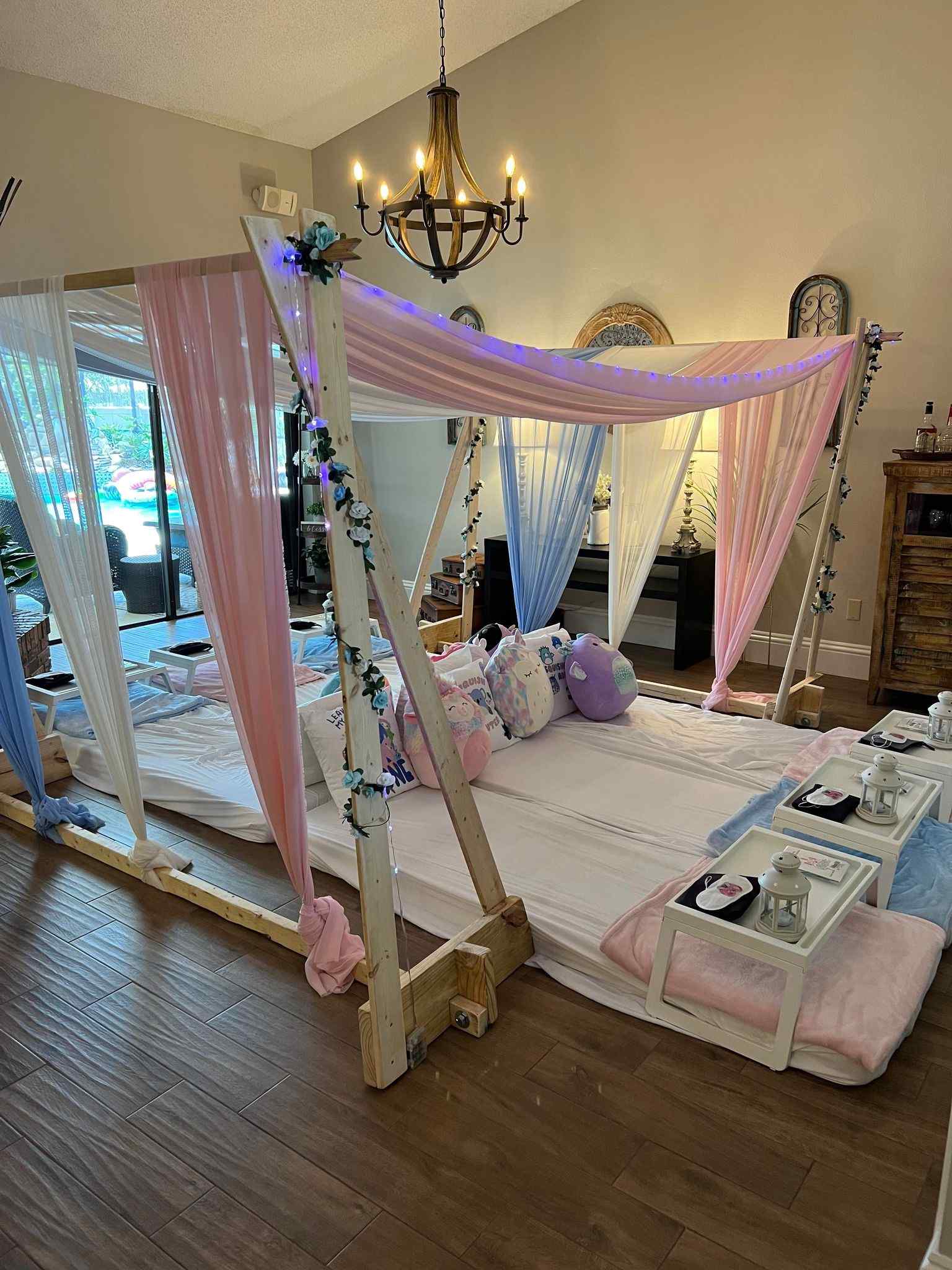 A cozy indoor canopy sleepover setup with draped pink and white curtains, decorative lights, and surrounding plush cushions.