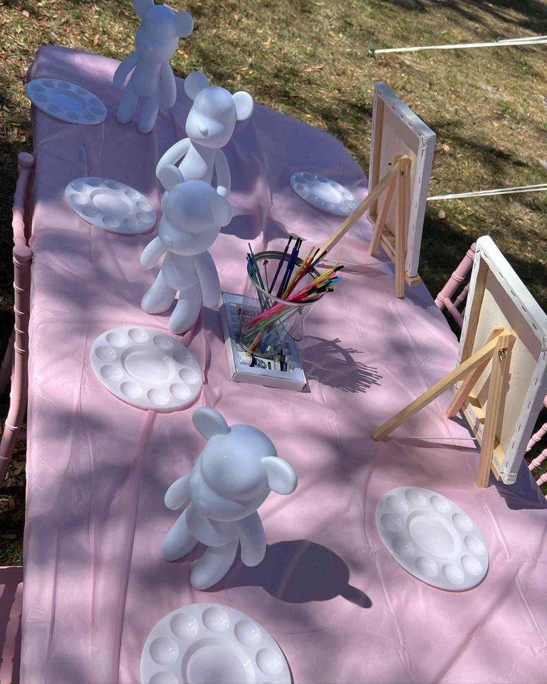 A creative outdoor paint station set up on a Pink Teddy Bear tablecloth featuring blank white figurines, palettes, and colorful markers ready for an art activity in a sunny setting.