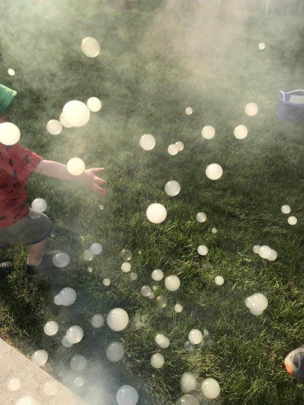 A child reaches out to catch bubbles floating in the air on a sunny day, surrounded by a haze from a Frogger F4 Bubble Fogger Rental and green grass.